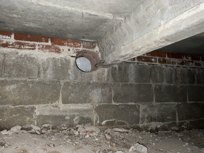 Hål tagit i källarens betongvägg nära en balk, exponerad armering synlig. Omgivande tegel och betong, dammig golvyta.