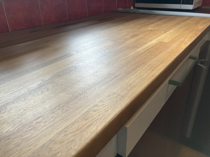 Köksskiva i trä behandlad med två lager hårdvax, med röd kakelvägg i bakgrunden och en del av en vit låda under.