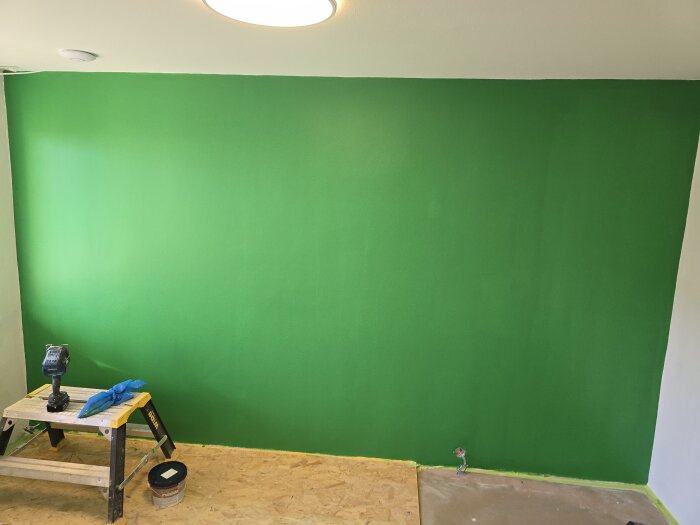 Nymålad grön vägg med stege, borrmaskin och färgburk framför. Golvtäckning delvis synlig, under pågående renovering. Taklampa lyser ovanifrån.