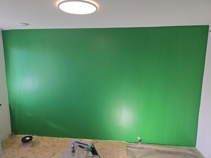 Nymålad grön vägg med jämn yta, verktyg och färgburk på golvet. Taklampa och snett dagsljus syns upptill i bilden.