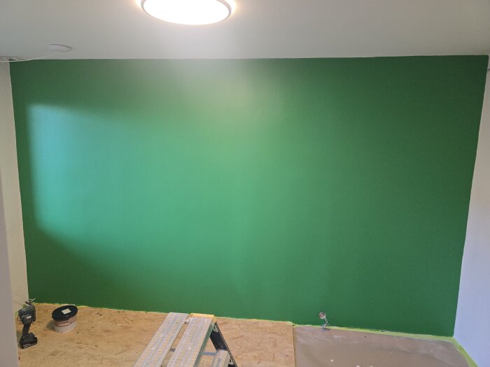 Nymålad grön vägg i ett rum, med en stege och färghink på golvet. Ett takljus lyser upp väggen.