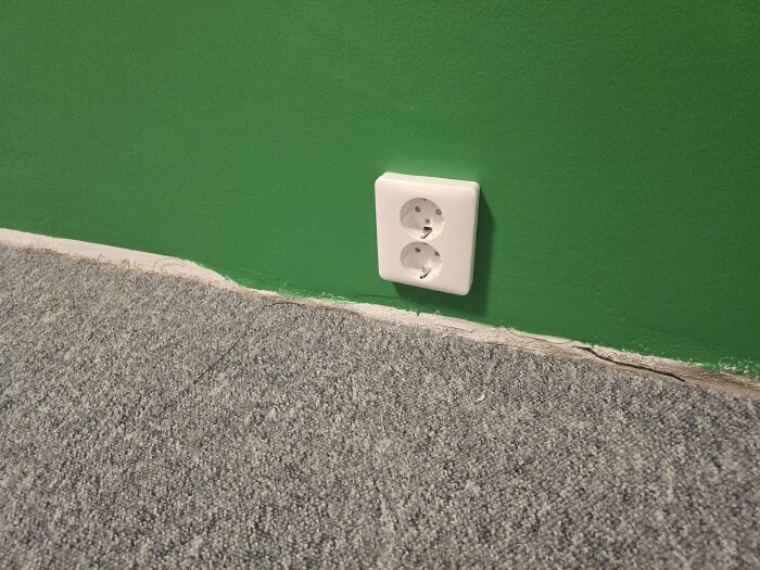Grönt nymålat rum med ett vitt dubbeluttag på väggen, synlig golvlist över grå matta.