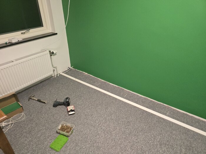 Ett rum under renovering med gröna väggar, grått golv, en golvlist, en borrmaskin, hammare och skruvar vid sidan av ett fönster och element.
