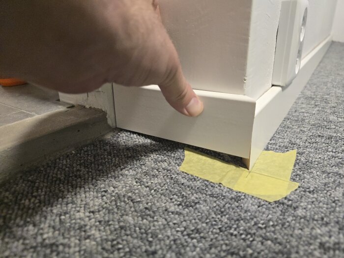 Hand som justerar snedkapad golvlist vid tröskel i barnrum, med tejp på heltäckningsmatta.