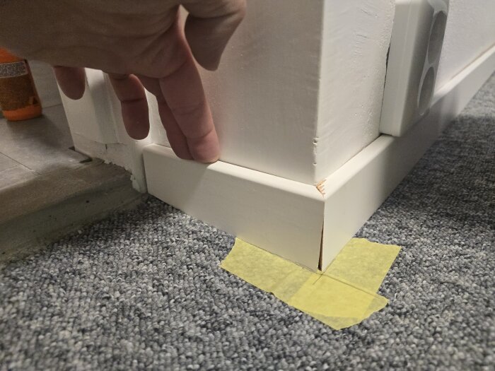 En hand visar en snedkapad vit list vid en vägg, med golvet täckt av grå matta och gult maskeringstejp på plats.