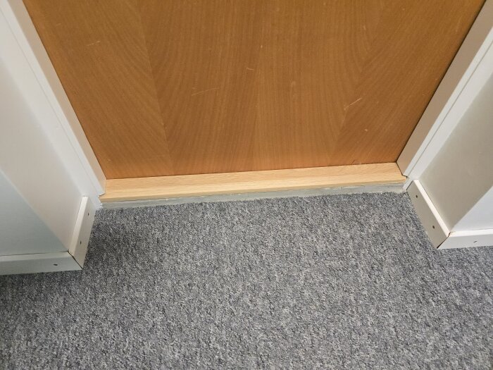 Nyinstallerad tröskel under dörr, omgiven av vit lister och grå heltäckningsmatta i barnrum.