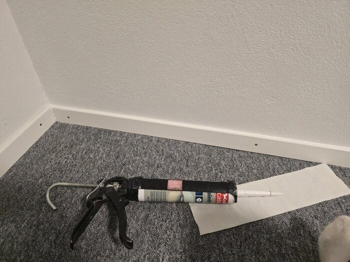 Fogpistol på golv bredvid nymonterad list i ett hörn, med skruvhål synliga och papper intill.