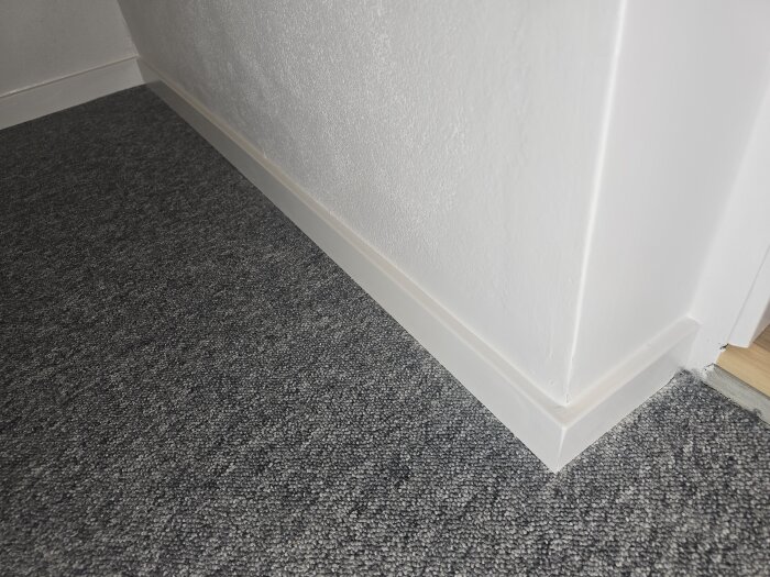 Vitt golvlister hörn på grått mattgolv, med jämn väggmålning. Förbättrat efter snedkapning och målning. Ny ljus tröskel syns delvis.