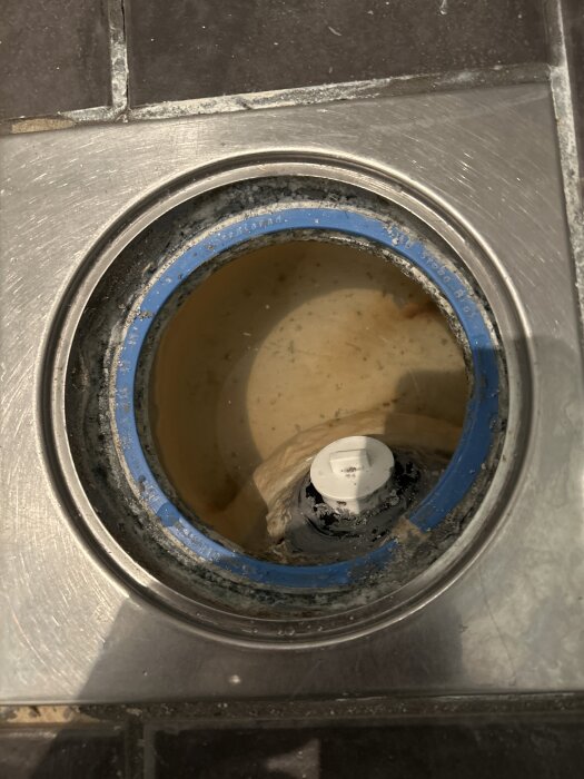 Gammal golvbrunn med gjutjärnsrenshål, omgiven av nytt golv. Vit propp synlig i mitten, omgärdad av blå kant.