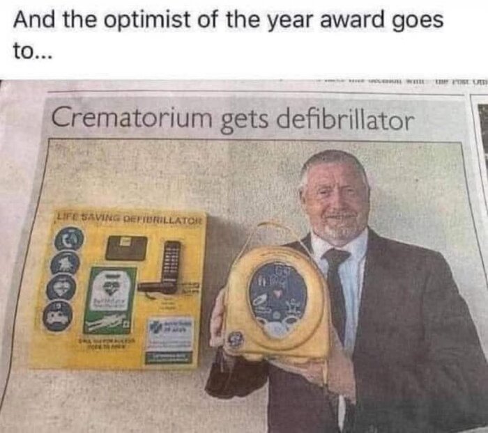 Tidningsbild med texten "Crematorium gets defibrillator". En man håller en defibrillator och står leende bredvid en skylt om livräddande utrustning.