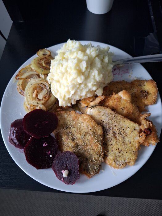 Panerad spätta med potatismos, stekt lök och skivade rödbetor på en vit tallrik.