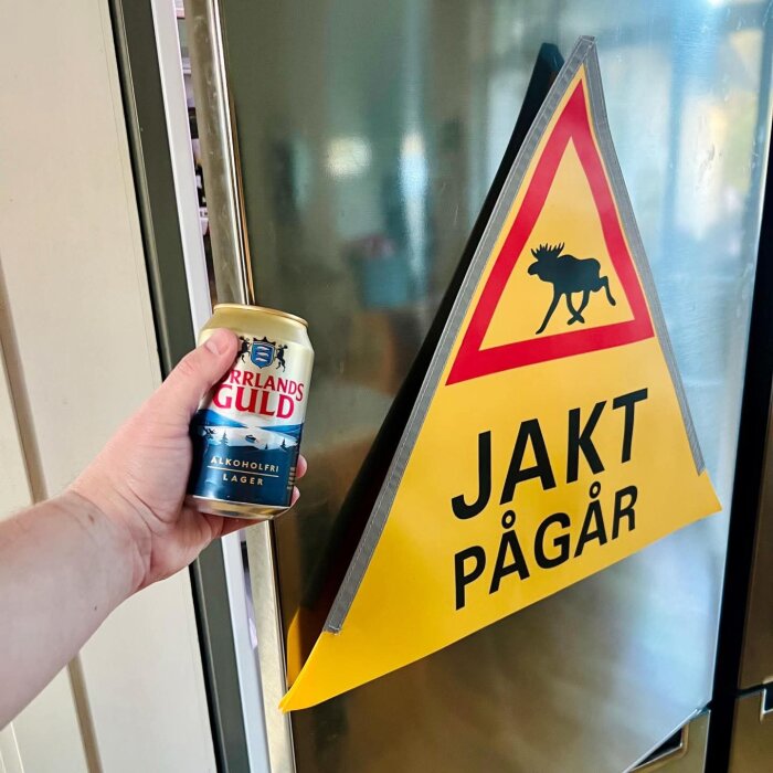 En hand håller en alkoholfri ölburk bredvid en gul skylt med texten "Jakt pågår" och en älg, monterad på ett kylskåp.