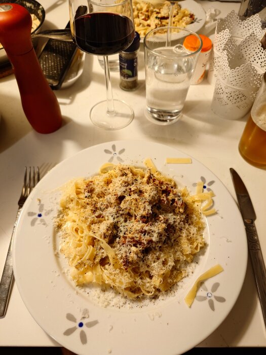Tallrik med pasta toppad med riven parmesan och svartpeppar, omgiven av glas vin och vatten, samt en pepparkvarn bredvid.