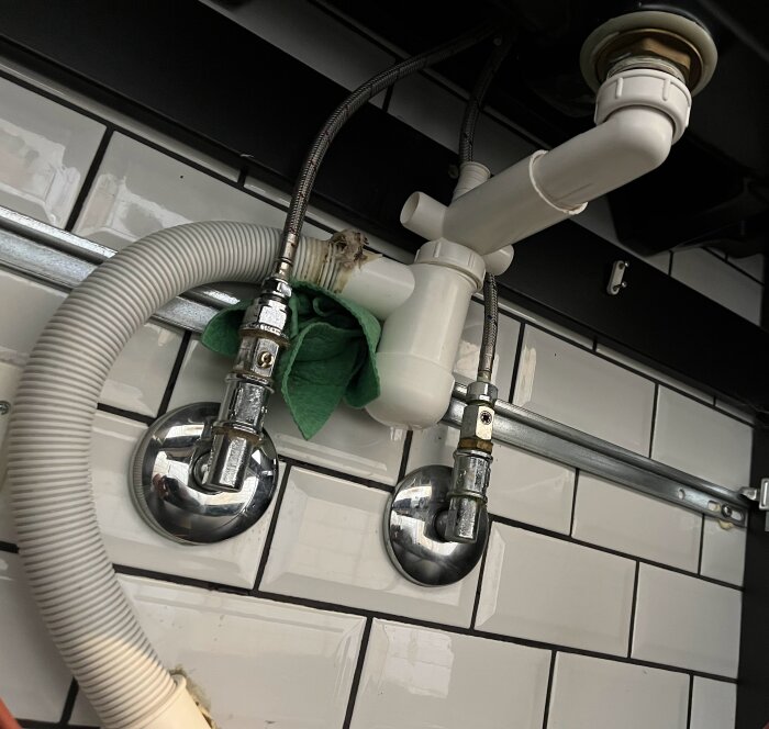 Läckande rör och vattenlås under ett handfat där en disktrasa är insatt vid övergången för att stoppa läckage, omgiven av vita kakelplattor.