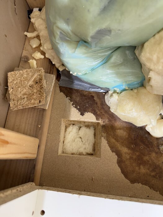 Öppet hål i träskiva med isolering under, omgiven av byggmaterial och plastisk massa.