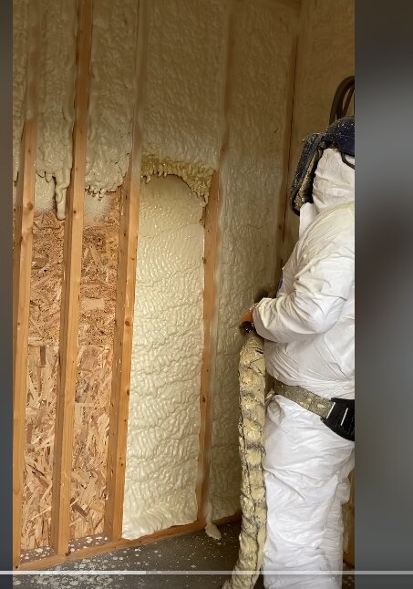 Person i skyddsdräkt applicerar isoleringsskum på väggar i ett rum, troligen polyuretanskum används för att isolera mellan trästolpar.