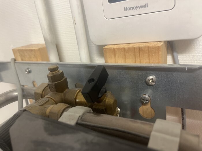 Ventilvred på fjärrvärmecentral, närbild av svart vred monterat på röranslutning under Honeywell-termostat på metallpanel.