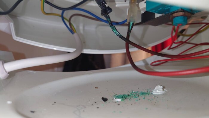 Elektriska kablar med ärgade och smälta delar inom ett värmesystem, med grön korrosion synlig på vit yta.