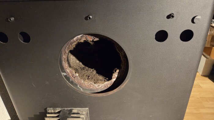 Öppning på baksidan av en kamin där en rund täckplåt har lossnat och fallit in i kaminens innandöme. Tre andra hål syns också på panelen.