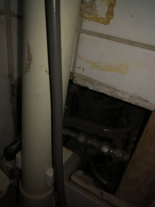 Rörsystem bakom tvättmaskin; synliga vattenrör och ett avloppsrör i ett litet utrymme med kaklade väggar och osäkrad öppning.