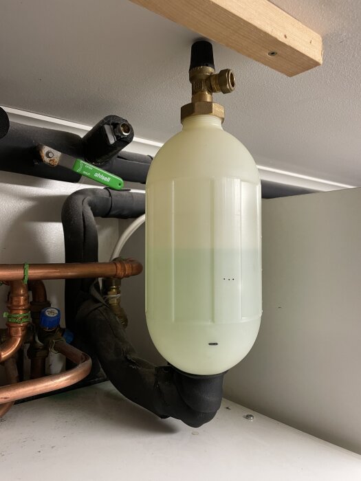 Expansionskärl för käldbärarvätska med påfylld 50/50-blandning, kopplat till VVS-system under ett tak med synliga kopparrör och ventiler.