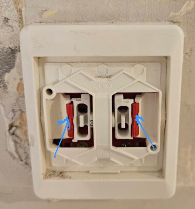 Väggmonterat vägguttag med två rödvita plastkomponenter och blå pilar som pekar mot dem.