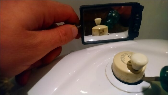 Hand som håller en spegel ovanför spolknappen på en Porsgrund-toalett från 2008, visar detalj på lockets fäste.