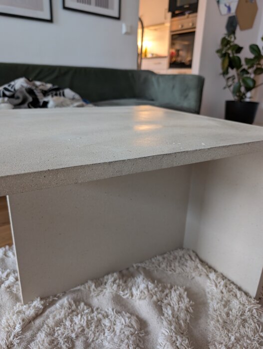 Bord med ljus yta, möjligtvis sprayad spånskiva, placerat på en vit matta. Bakgrund: mörkgrön soffa, växt och kök.