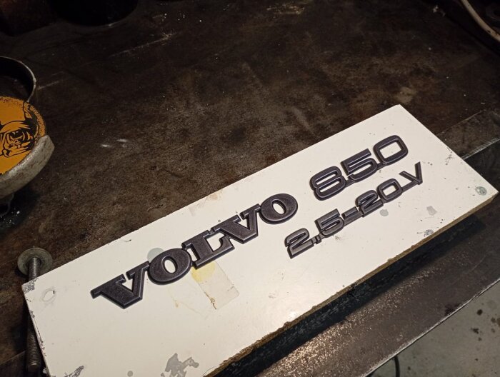 Svarta Volvo 850 2.5-20V emblem på vit bakgrund, placerade på arbetsbänk i garage.