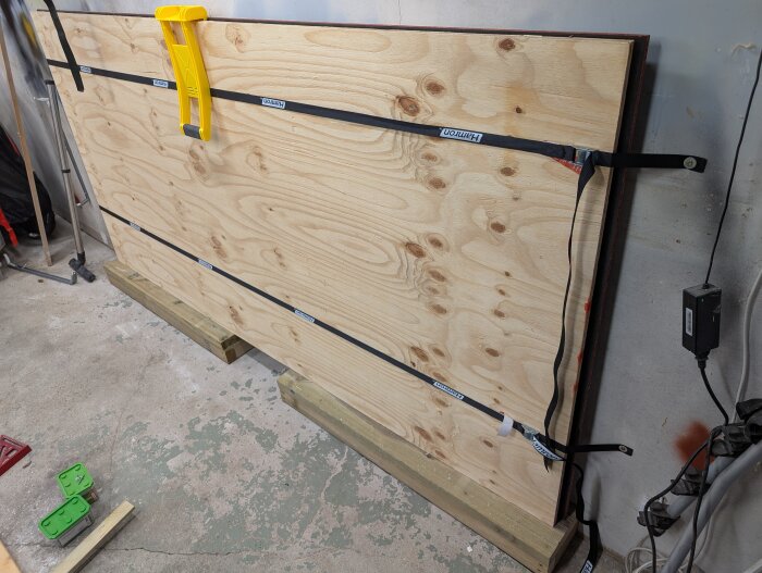 Förvaring av skivmaterial i ett garage med fuktigt golv, skivorna hålls på plats med remmar och en gul skivlyftshållare syns.