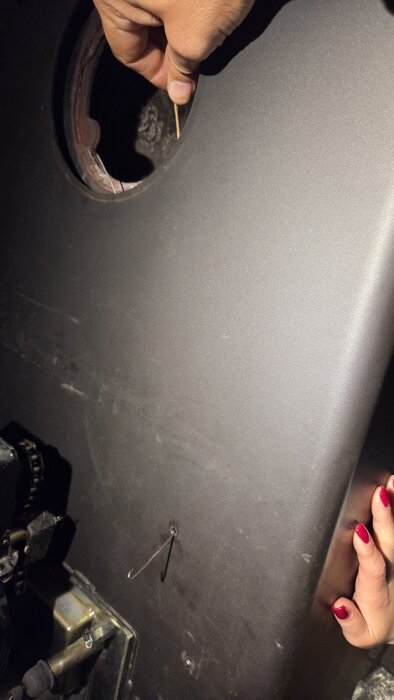 Händer justerar en panel med ett borrat hål och en tråd, del av en kamin eller pelletsmatare.