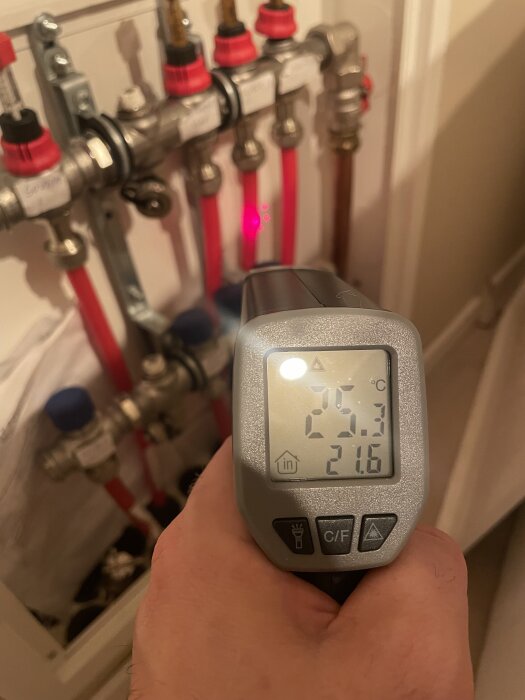 En hand håller en IR-termometer som visar temperatur vid en golvvärmefördelare med rör, ventiler och flödesmätare.