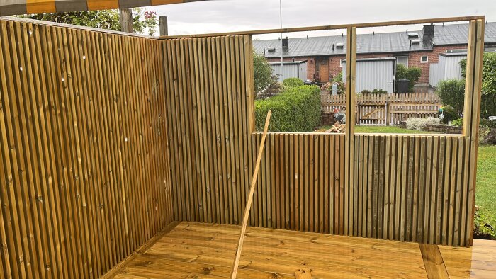 Trästaket under konstruktion, plan för plåt ovanpå och eventuell dold ljusslinga under kant för belysning av ribborna, trädgård i bakgrunden.