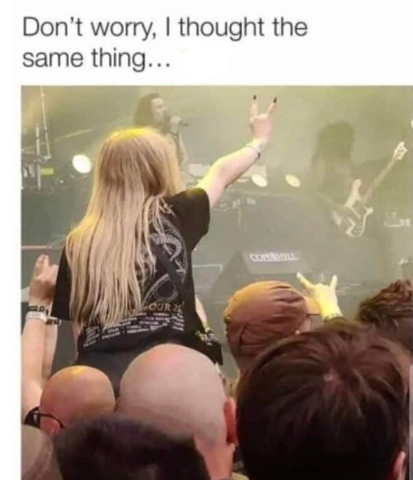 En person med långt hår som håller upp rocktecken med handen på en konsert, står ovanpå andras axlar.