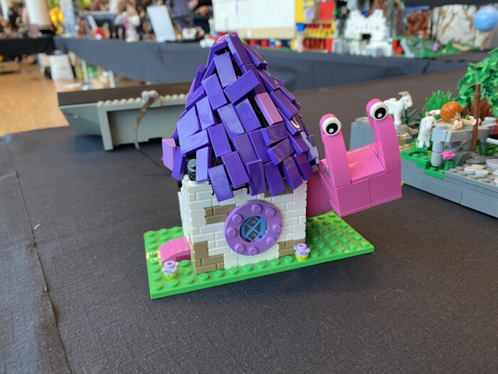 Lego-snigelhus med lila tak och rosa snigelhuvud med ögon, uppvisning i en hall med andra Lego-kreationer i bakgrunden.