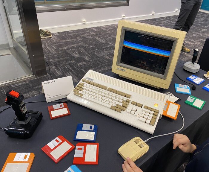 En Amiga 1200-dator med tangentbord, mus och skärm, omgiven av färgglada disketter och en joystick på ett bord.
