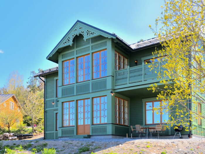 Grönt tvåvåningshus med utsmyckade fönster och en uteplats, omgiven av vårblommande träd och gräsmatta.