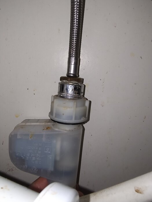 Plastmutter och metallrörsdel på diskmaskinens vattenanslutning, med en genomskinlig plastbehållare i närheten.