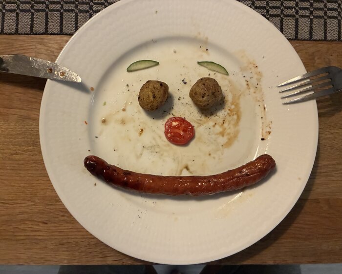 Tallrik med matrester arrangerade som ett ansikte, med gurka som ögonbryn, köttbullar som ögon, tomat som näsa och korv som mun.