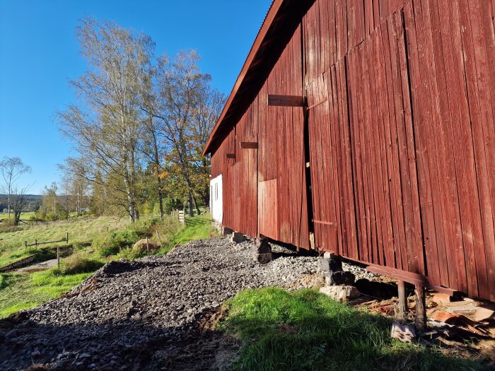 Röd träloge under renovering med synliga grundstenar och fyllnadsmaterial. Platsen är delvis grästäckt med några träd i bakgrunden.