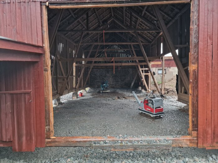 Renovering påbörjad i lada med öppen dörröppning, där golvet täcks av grus och en markvibrator står redo för användning.