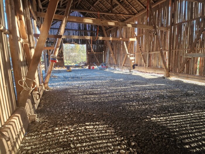 Inuti en loge under renovering, med ett golv täckt av stenar och grus, synliga träbalkar i taket och väggarna. Solens ljus strömmar in.