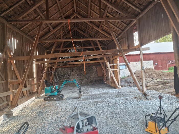 En loge under renovering med en grävmaskin och packad grusgrund, omgiven av träbjälkar.