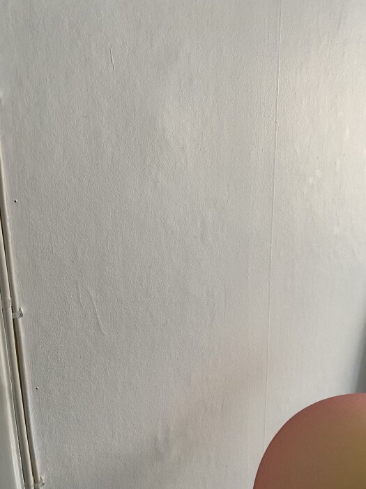 En vägg med ojämnheter och små skador, eventuellt behöver bredspacklas; en sladd ses på vänster sida och något orange i nedre hörnet.