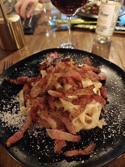 Flamberad carbonara serverad på en svart tallrik, med knaperstekt bacon och parmesan, på ett trämatsalsbord med ett glas rödvin i bakgrunden.