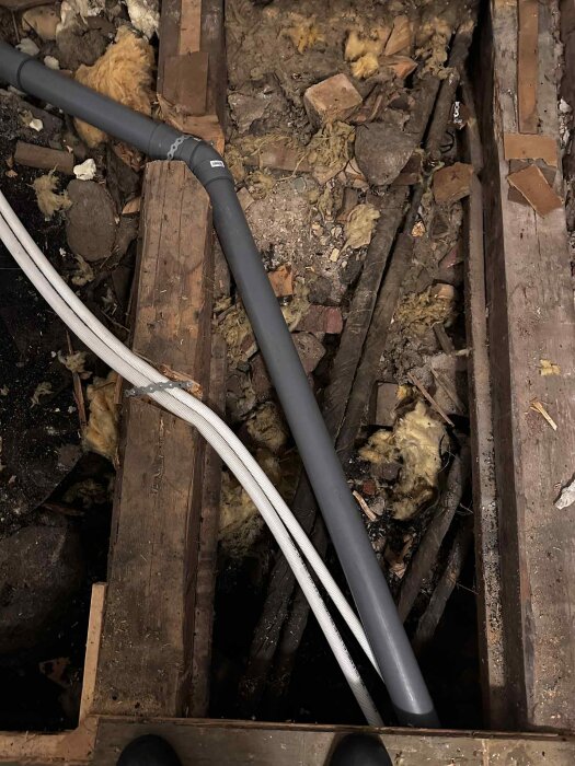 Bilden visar en gammal krypgrund med nya avloppsrör och synligt bjälklag. Det finns sten och isoleringsmaterial mellan bjälkarna.