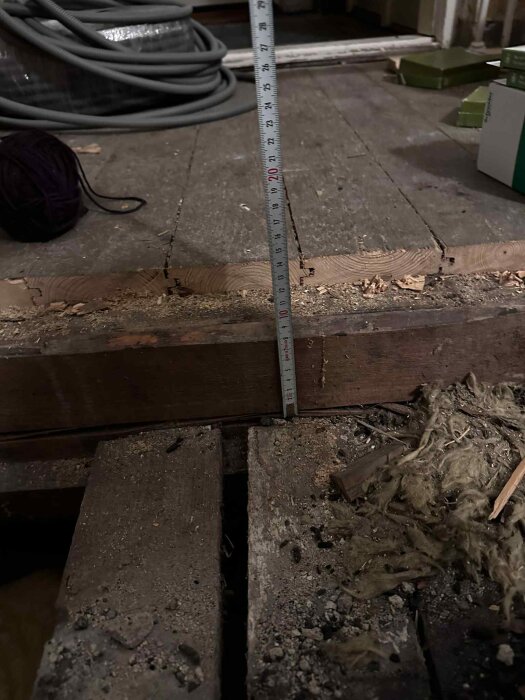 Golv uppbrutet för renovering, visar mätskalor och träbjälkar. Trasig isolering och smuts syns. Bakgrund av rör och verktyg.