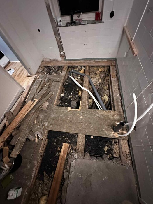 Öppet gammalt köksgolv med synliga bjälkar, rör och isolering. Rester av krossad koks och träspill syns, samt betong och trägolv.