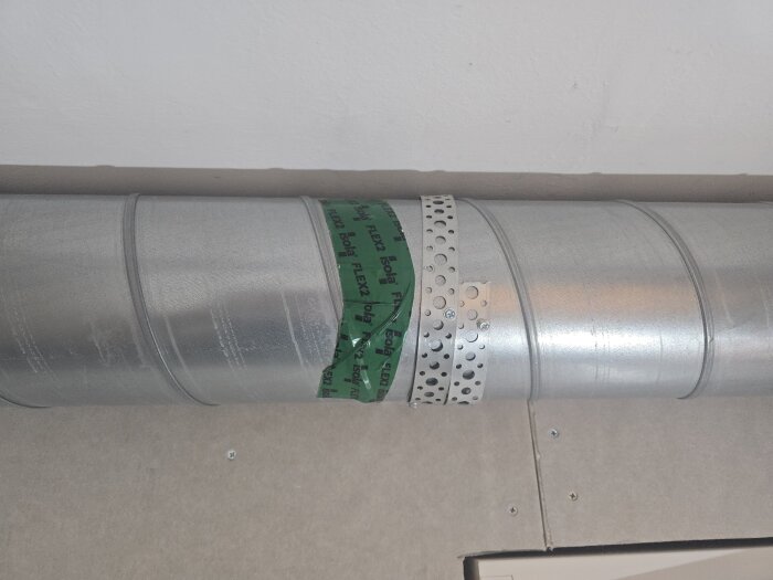 Ventilationsrör i metall med grönt tejp och perforerat metallband för att täta och fästa rör vid byggrenovering. Rör löper längs ett tak.