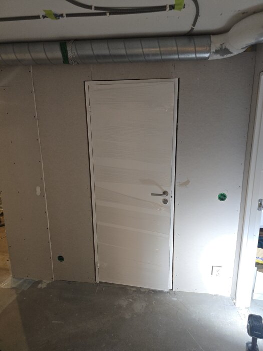 Vägg gipsad med dörr monterad i källarkorridor. Spirorör och gröntejp syns i taket, eluttag och takdosor förbereds. Arbetsverktyg på golvet.